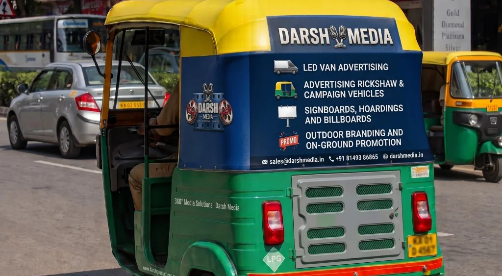 A green and yellow auto-rickshaw in India with a Darsh Media advertising wrap listing LED van, rickshaw, hoarding and on-ground promotion services, driving down a city street